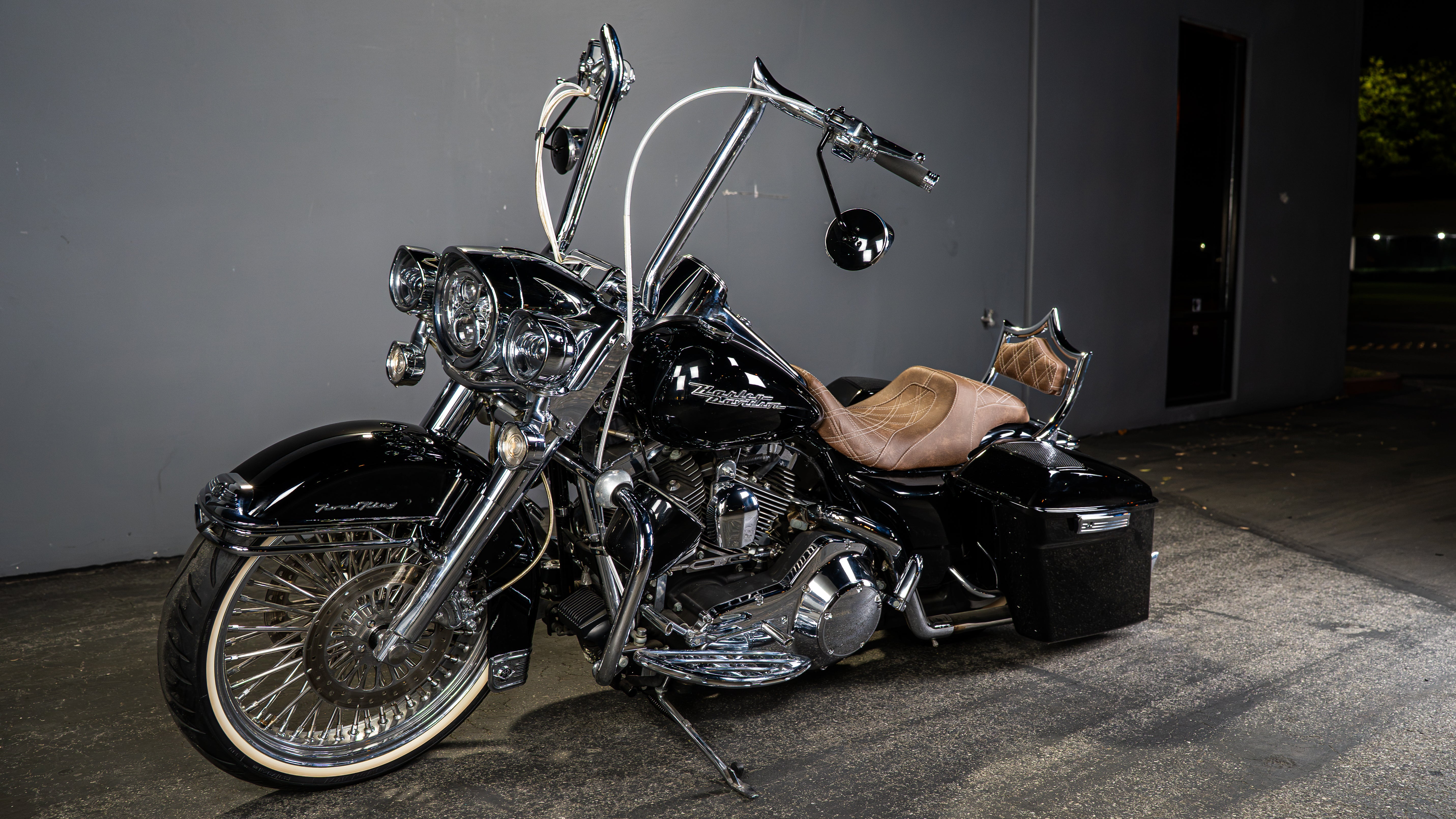 A black cruiser motorcycle with chrome details, a tan leather seat, and black saddlebags, parked against a dark gray wall at night.